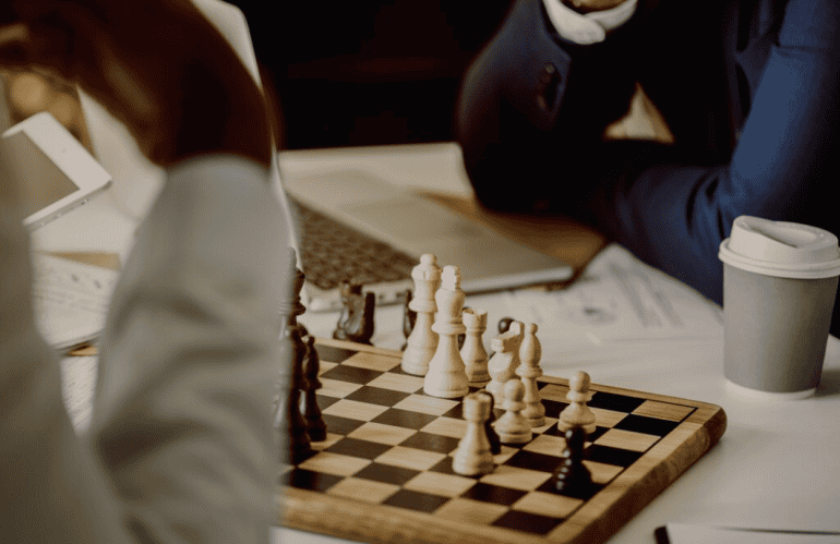 Two individuals engrossed in a chess game with a laptop and a coffee cup nearby